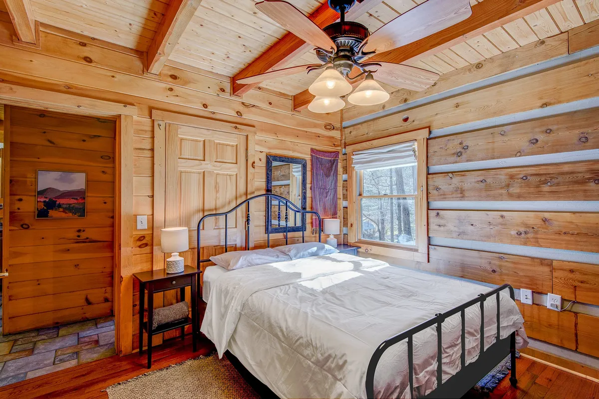 This image showcases a cozy queen bed set against wooden walls, with a window allowing natural light to stream in. The ceiling fan and overall setup evoke a calm, restful atmosphere ideal for unwinding after a day in the mountains.