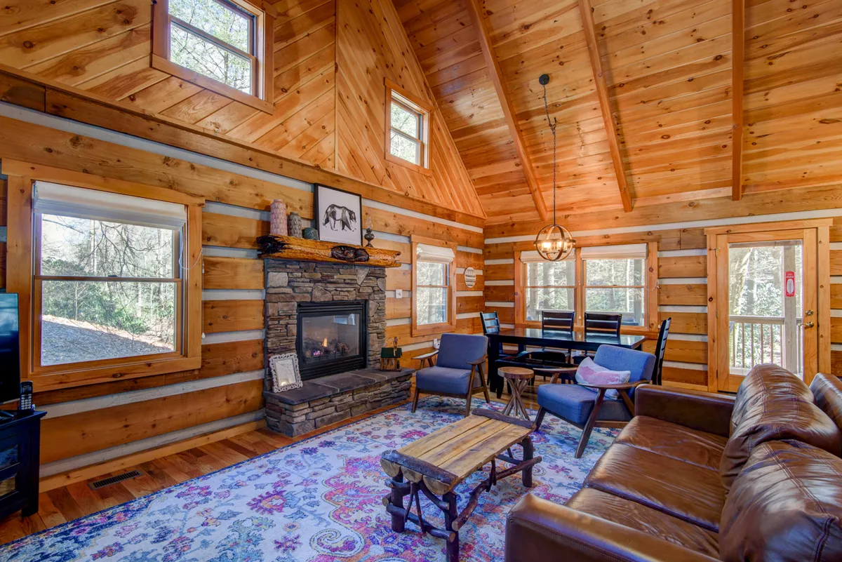 A beautifully appointed family room with a stone fireplace, wooden beams, a colorful rug, a dining table, and ample seating. The inviting ambiance is perfect for relaxing, dining, and playing games by the fire.