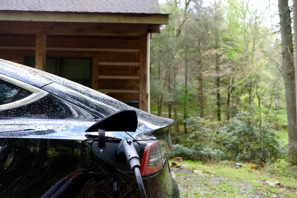 An electric vehicle is parked by a log cabin and connected to a charging station, surrounded by green woods. The scene is calm and eco-friendly, demonstrating the property's commitment to sustainability for modern travelers.