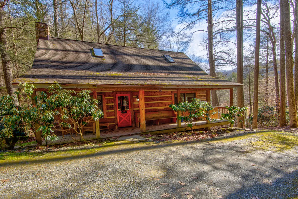 A charming log cabin with a striking red front door nestled beneath towering Hemlock trees, surrounded by lush forest. The inviting front porch looks out over the tranquil yard, perfect for relaxing, watching sunsets, or gathering around a campfire. The setting promises peace, privacy, and a deep connection to nature.