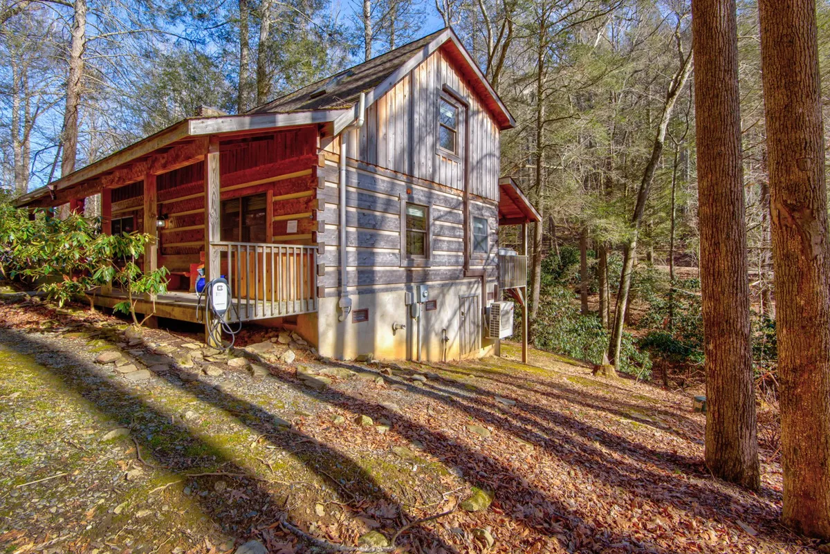 The wooden cabin is nestled among tall trees, with a cozy porch area visible out front. The surrounding greenery and peaceful ambiance evoke the tranquility of the mountain location.