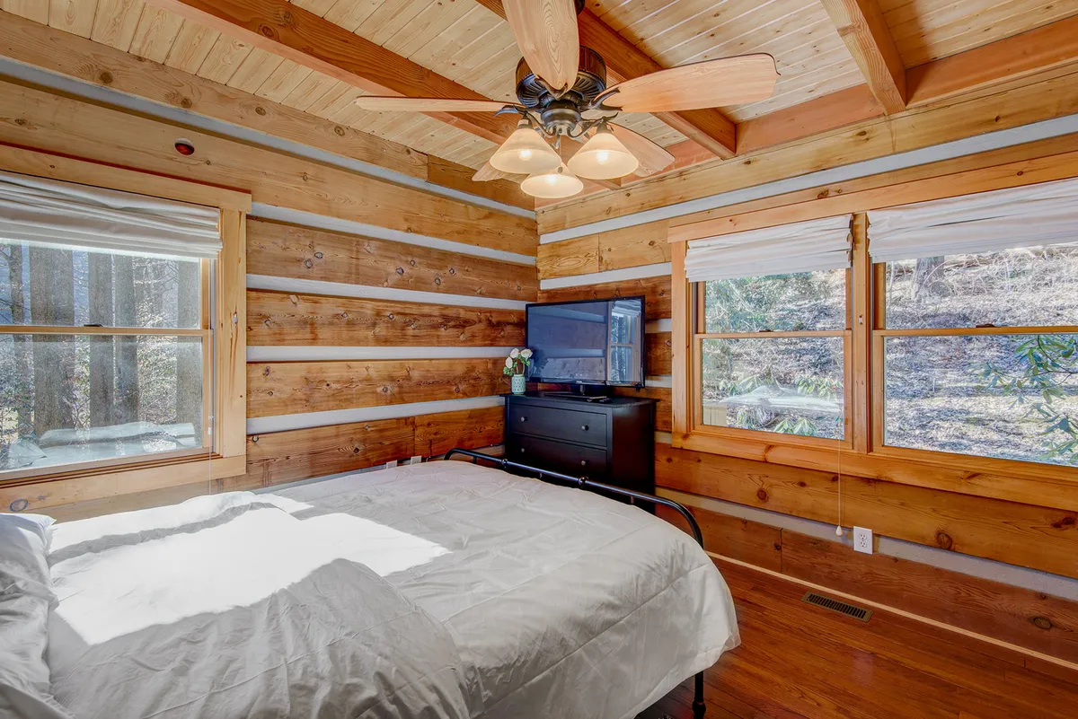 A tranquil bedroom featuring a queen bed with crisp white linens, wooden walls, a ceiling fan, and a television. Sunlight filters in, creating a bright and comfortable retreat for restful nights.