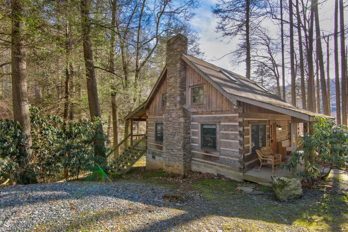 The log cabin's structure is surrounded by tall trees and features a spacious front porch with a chair. The natural setting highlights the property's proximity to nature trails and emphasizes its peaceful environment.