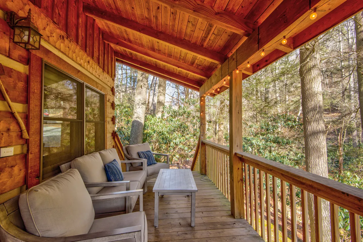 A spacious wooden deck adorned with string lights and rocking chairs overlooks a lush natural setting. The wooden railing blends seamlessly with the surrounding trees, evoking a sense of deep tranquility. The inviting outdoor space is perfect for relaxing, reading, or simply soaking up the gentle sounds of the nearby stream.