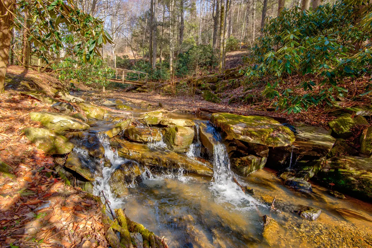 A tranquil exterior scene featuring a flowing waterfall framed by rugged rock formations and lush green surroundings. The gentle movement of water, dappled sunlight, and wild greenery create a serene, immersive natural setting ideal for quiet reflection or family exploration.