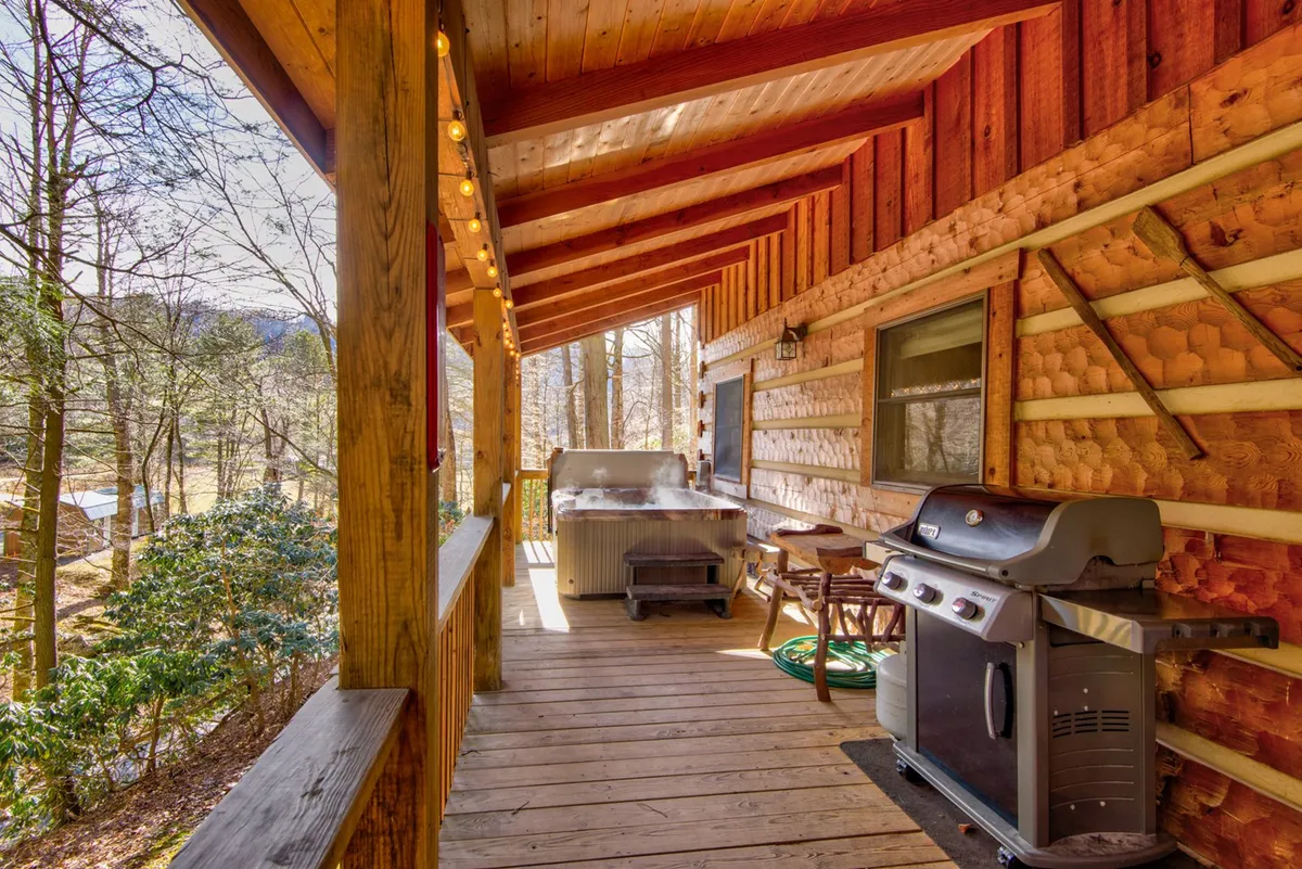 A cozy outdoor living area featuring a wooden deck with both a hot tub and a gas barbecue grill. The deck is accented by string lights and natural wood railings, creating a warm and inviting space ideal for barbecuing, soaking, and socializing amid nature.
