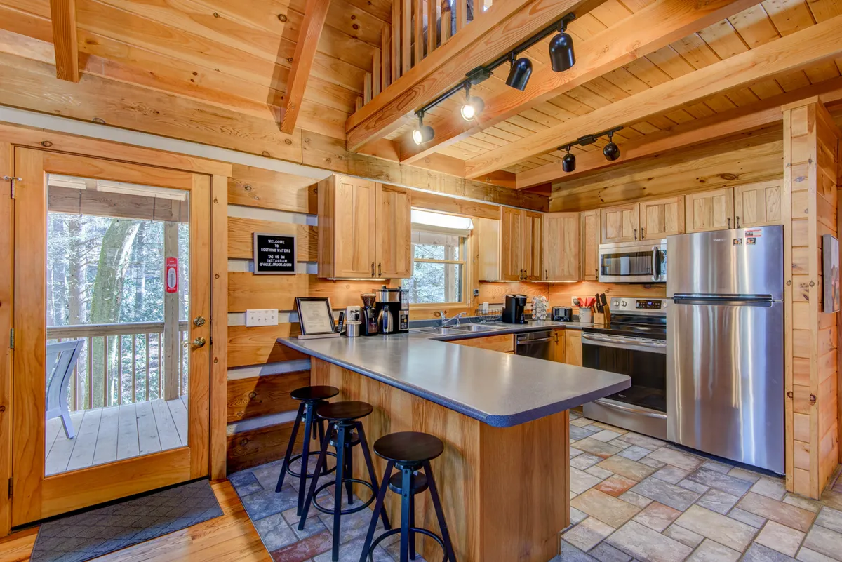 A modern rustic kitchen with a spacious island, stainless steel appliances, warm wooden cabinetry, and large windows that bathe the room in natural light. The setup is ideal for cooking, entertaining, and family meals.