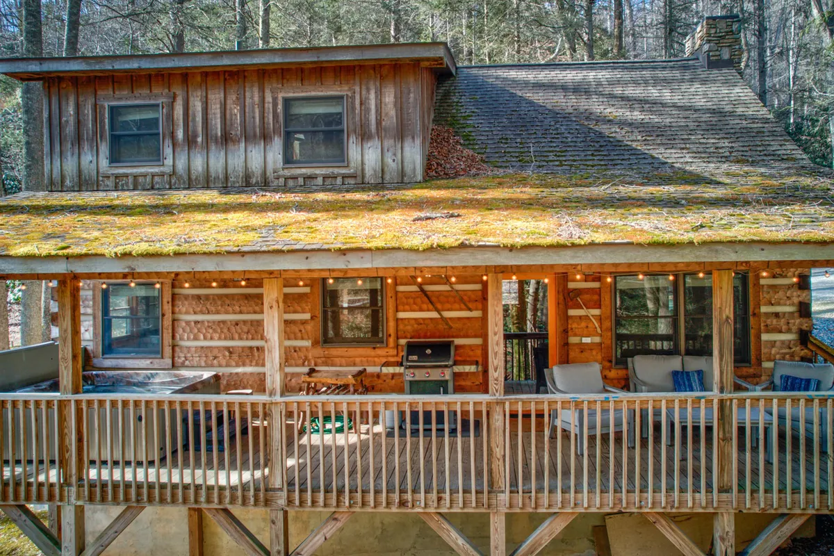 This image showcases the log cabin's exterior with a moss-covered roof, a spacious deck featuring a seating area and grill, and lush trees all around. The deck invites alfresco dining and relaxation, connecting guests to the outdoors in any season.